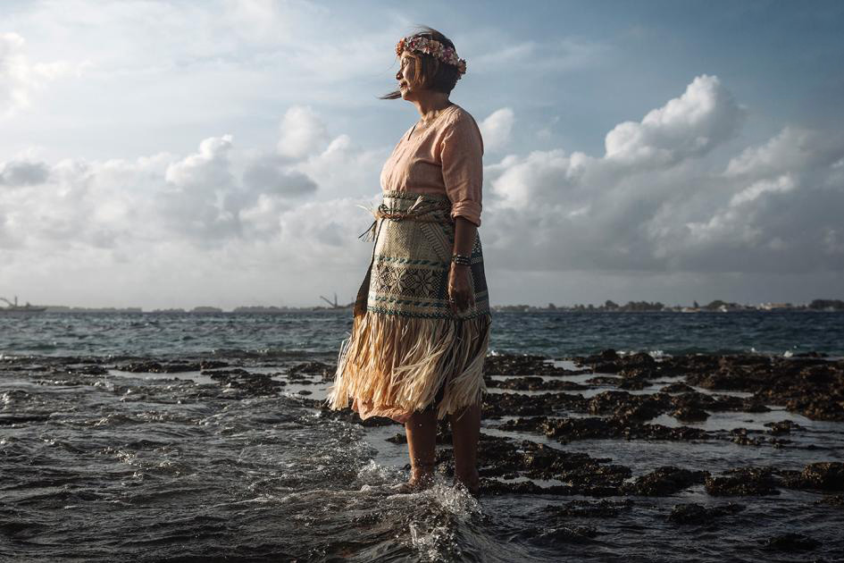 A woman in a traditional outfit stands with her feet in the ocean