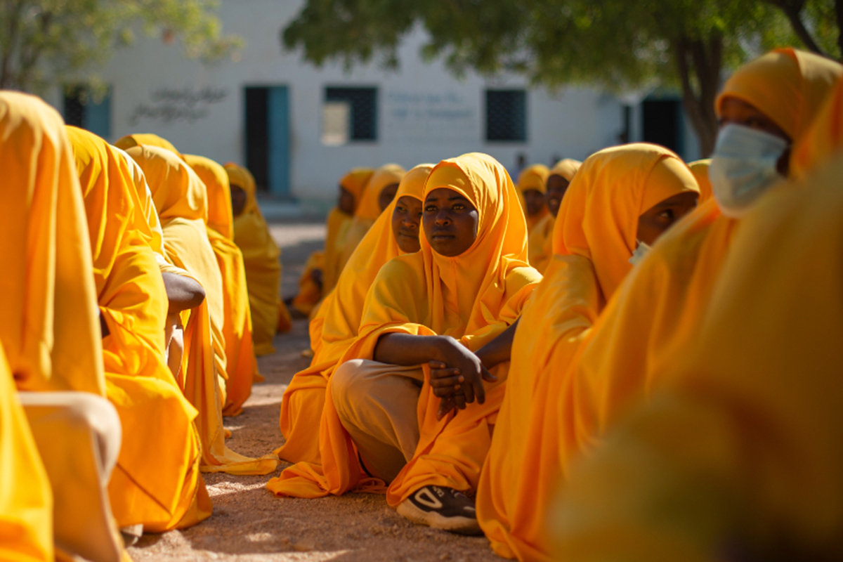 Girls in orange hijabs sit lined up outdoors.