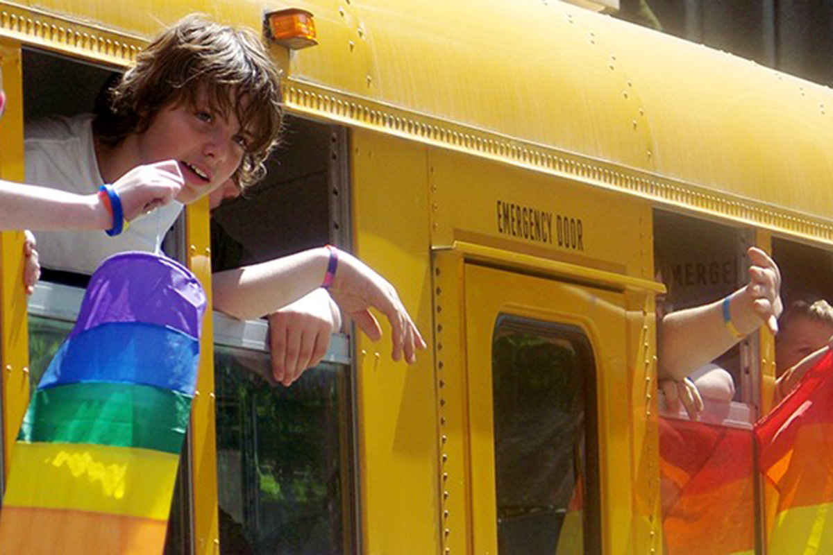 Students hold out rainbow flags from a school bus.